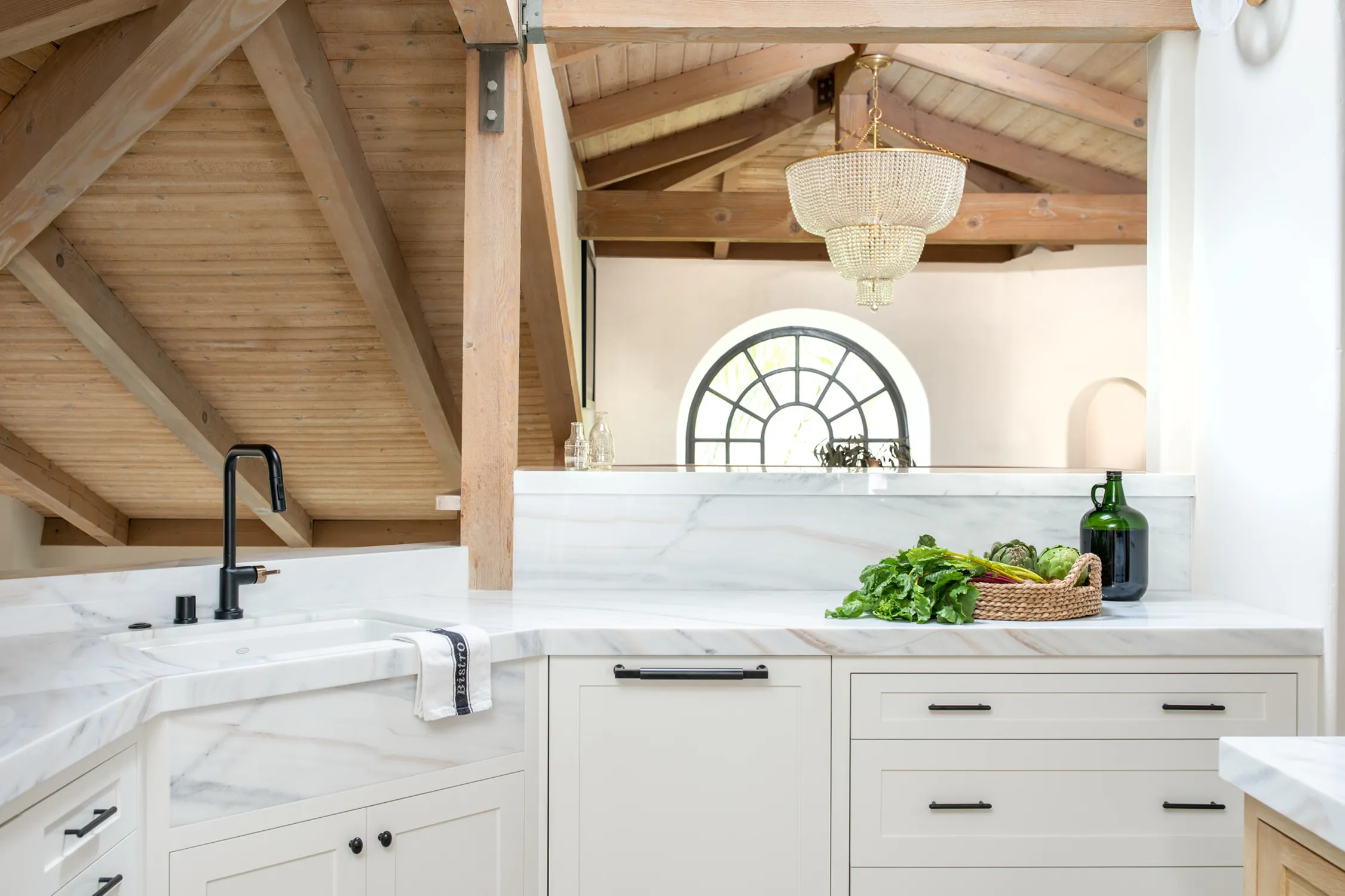 White kitchen beneath exposed wood beams and arched window